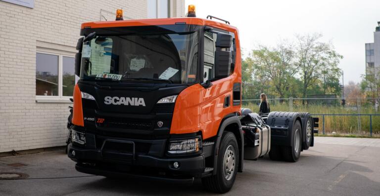 Oranžen tovornjak Scania pred šolskim vhodom.